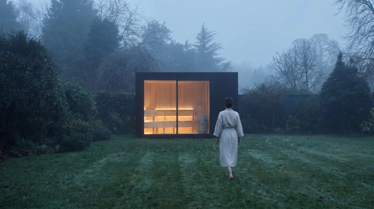 A lit NOOK Sauna in a misty garden, with someone in a bathrobe walking towards it